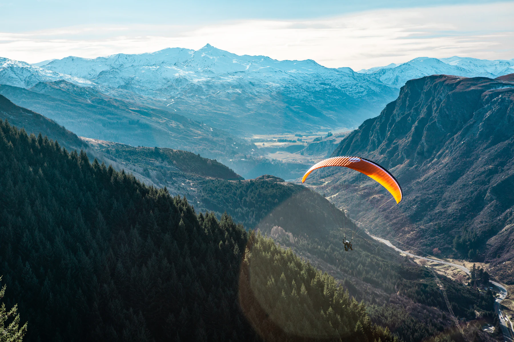 An orange paraglider soaring above a forested valley and snow-covered mountains near Queenstown